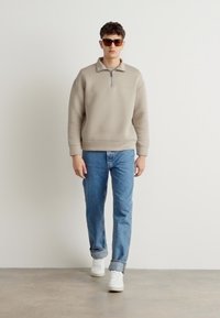 Felpa beige con colletto a zip a un quarto, abbinata a jeans azzurri chiari e sneakers bianche. La felpa presenta una texture liscia.
