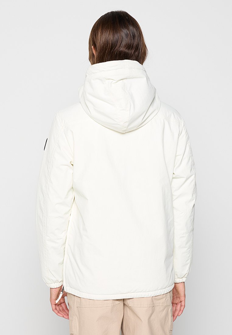 Person mit langen braunen Haaren, von hinten gesehen, trägt eine weiße Kapuzenjacke und beige Hosen vor einem schlichten Hintergrund.