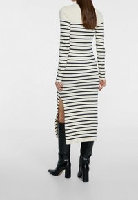 Abito in maglia a righe crema e navy con maniche lunghe, spacco laterale e design aderente, abbinato a stivali al ginocchio lucidi neri.