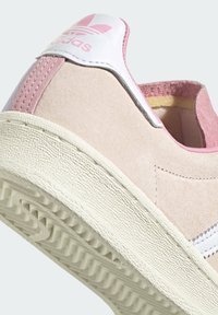 Bledě růžové semišové tenisky s bílými akcenty, s texturovanou gumovou podrážkou a růžovým poutkem na patě. Logo Adidas na zadním panelu.