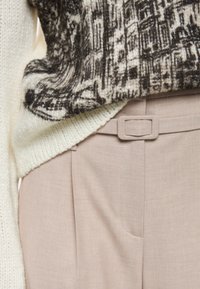 Knitted sweater with a cream and brown pattern layered over light beige tailored trousers, featuring a textured finish and a belt loop accent.