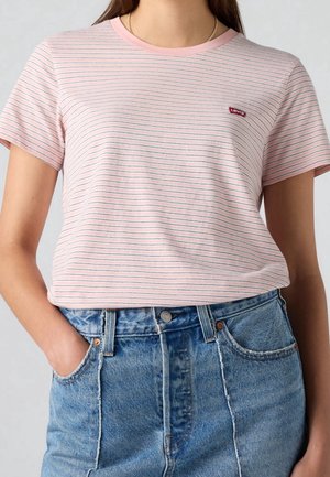 Ung person iført en lyserød Levi's t-shirt med tynde striber, stoppet ned i højtaljede blå cowboybukser, med den ene hånd i lommen.