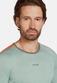 Ljusgrön långärmad skjorta med grå detaljer och klara orange axlar, med en logotyp på bröstet. Mjuk, texturerad tyg.