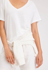 Maglietta bianca a collo a V in un tessuto leggero con maniche corte, abbinata a un pantalone chiaro; un maglione bianco è annodato in vita.