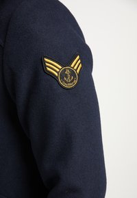 Rameno námořnické vlněné bundy se zlatou křídlatou insignií a kulatou nášivkou s kotvou a nápisem "DREMASTER". Měkká textura, strukturovaný střih.