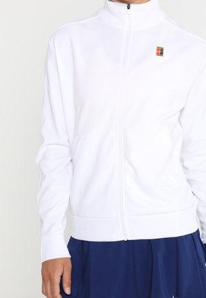 Person wearing a white zip-up jacket with a small colorful logo on the chest and a navy blue skirt or pants.