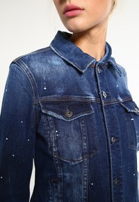 Jeansjacka i mörkblått, med två bröstfickor, metallknappar och utspridda vita färgfläckar. Klassisk kragsdesign.