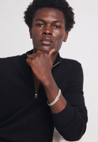 Sweat à zip noir avec des manches côtelées, porté par une personne avec un bracelet en argent, présentant un accent rouge et bleu. Texture de peau lisse.