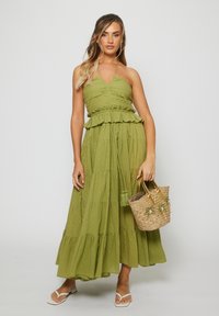 Woman wearing a sleeveless olive green tiered maxi dress holding a woven straw handbag with palm tree embellishments.