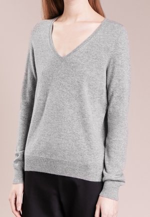 Femme portant un pull col en V gris clair à manches longues et un pantalon noir, debout devant un fond beige uni.