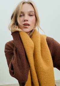 Mladá žena s voľnými blond vlasmi, oblečená v hnedom rebrovanom svetri a hrubej horčicovo žltej šále na jednofarebnom pozadí.