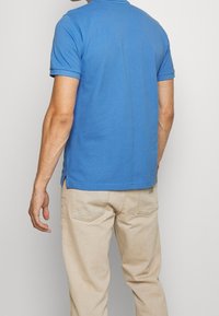 Modrá bavlněná polokošile s krátkým rukávem, s mírně delším zadním lemem a bočními rozparky, v kombinaci s béžovými kalhotami.