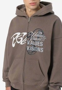 Braune Kapuzenjacke mit Reißverschluss aus weichem Stoff, mit Fronttaschen. Aufgedruckter Text enthält "Values" und "Visions" in verschiedenen Schriftarten und Farben.