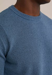 Suéter de punto azul con una superficie texturizada, cuello redondo acanalado y puños ajustados. La tela parece suave y ligeramente texturada.