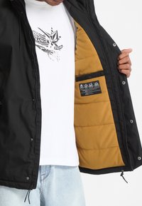 Personne portant une veste noire avec une doublure beige, la tenant ouverte pour montrer l'étiquette intérieure, portant un t-shirt blanc graphique et un jean bleu clair.
