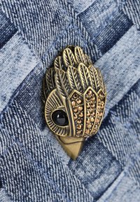 Mosazná brož ve tvaru ptáka s detaily listů, ozdobená bronzovými kamínky a černým akcentem, na texturovaném modrém denimovém pozadí.