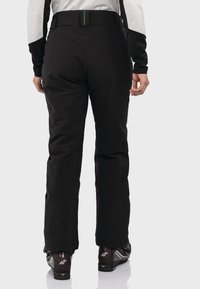 Schwarze Skihosen mit hohem Bund, mit grünem Stickdetail, und Schneestiefeln an den Füßen. Glatte Textur und taillierte Passform.