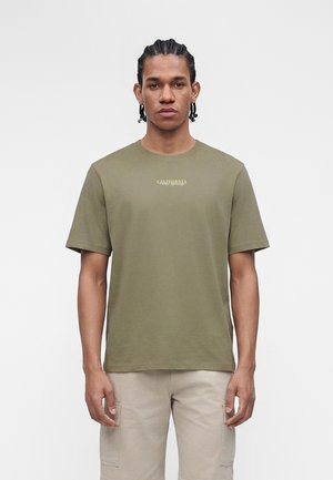 Jeune homme aux cheveux tressés portant un t-shirt vert olive "California" et un short cargo beige, debout devant un fond clair uni.