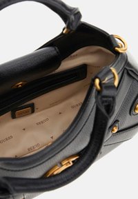 Sac à main en cuir noir avec des accessoires dorés, ouverture sur le dessus révélant un intérieur en tissu beige avec des logos Guess imprimés et une poche intérieure zippée.