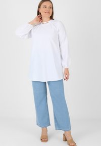 White long-sleeve top with a round neckline and gold-tone hardware detail, paired with light blue wide-leg jeans and tan block-heel sandals.