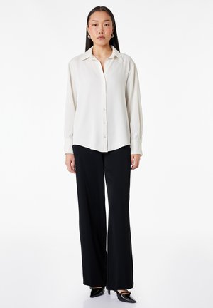 Frau steht, trägt ein weißes, langärmeliges Hemd mit Knöpfen, schwarze weitbeinige Hosen und schwarze spitze High Heels vor weißem Hintergrund.