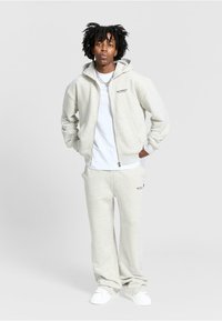 Light grey zip-up hoodie with front pockets and logo, paired with matching sweatpants. White t-shirt underneath. Smooth texture, casual fit.