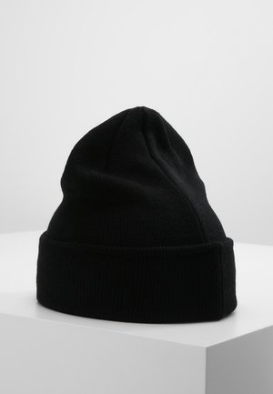 Bonnet noir en tricot avec un revers plié posé sur une surface blanche devant un fond gris clair uni.