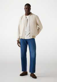 Licht beige bomberjack met verticale textuur, ritsluiting en geribde cuffs, gecombineerd met blauwe rechte spijkerbroek en bruine moccasinschoenen.