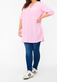 Camicia rosa a bottoni con scollatura rotonda e due tasche frontali, abbinata a jeans skinny blu scuro e sneakers bianche con strisce nere.