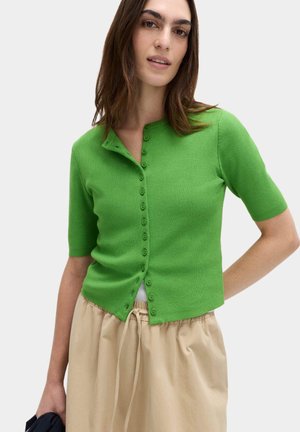 Frau trägt eine leuchtend grüne, geknöpfte Kurzarm-Strickjacke und beige Hosen mit Kordelzug, steht vor einem schlichten hellen Hintergrund.