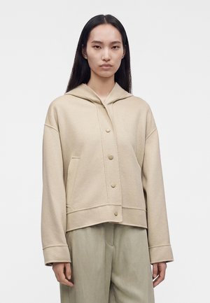 Jeune femme aux longs cheveux foncés portant une veste beige à capuche boutonnée et un pantalon ample de couleur claire sur un fond uni.