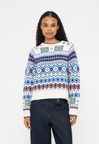 Maglione bianco lavorato a maglia con disegni a motivo in blu, verde e marrone; scollatura rotonda e polsini a coste; indossato con jeans scuri.