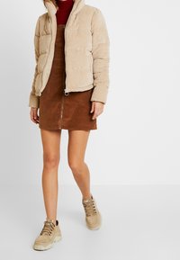 Beige fløjlspufferjakke med høj krave, parret med en brun fløjlskjole med frontlynlås. Fodtøj inkluderer beige chunky sneakers.