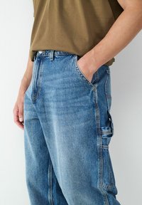 Pantalons cargo en denim bleu clair avec une coupe décontractée. Dotés de poches latérales et de coutures visibles. Assortis à une chemise à manches courtes marron.