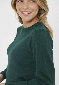 Zelený pletený sveter s rebrovaným okrúhlym výstrihom a balónovými rukávmi. Materiál vykazuje jemnú trblietavú textúru po celom povrchu.