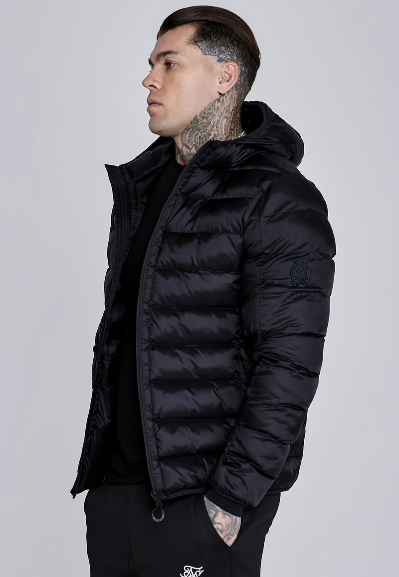 Chaqueta de plumas negra con capucha, diseño acolchado y cierre con cremallera. El material tiene una textura suave y brillante con un parche de logo en la manga.