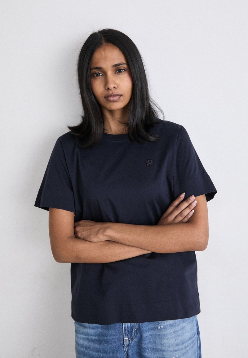 Navyblaue Baumwoll-T-Shirt mit kurzen Ärmeln und Rundhalsausschnitt; verfügt über ein kleines, tonales Logo auf der Brust. Trägt man mit blauen Jeans.