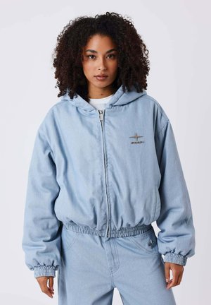 Femme aux cheveux bouclés portant un sweat à capuche bleu clair zippé et un pantalon assorti, debout devant un fond blanc uni.