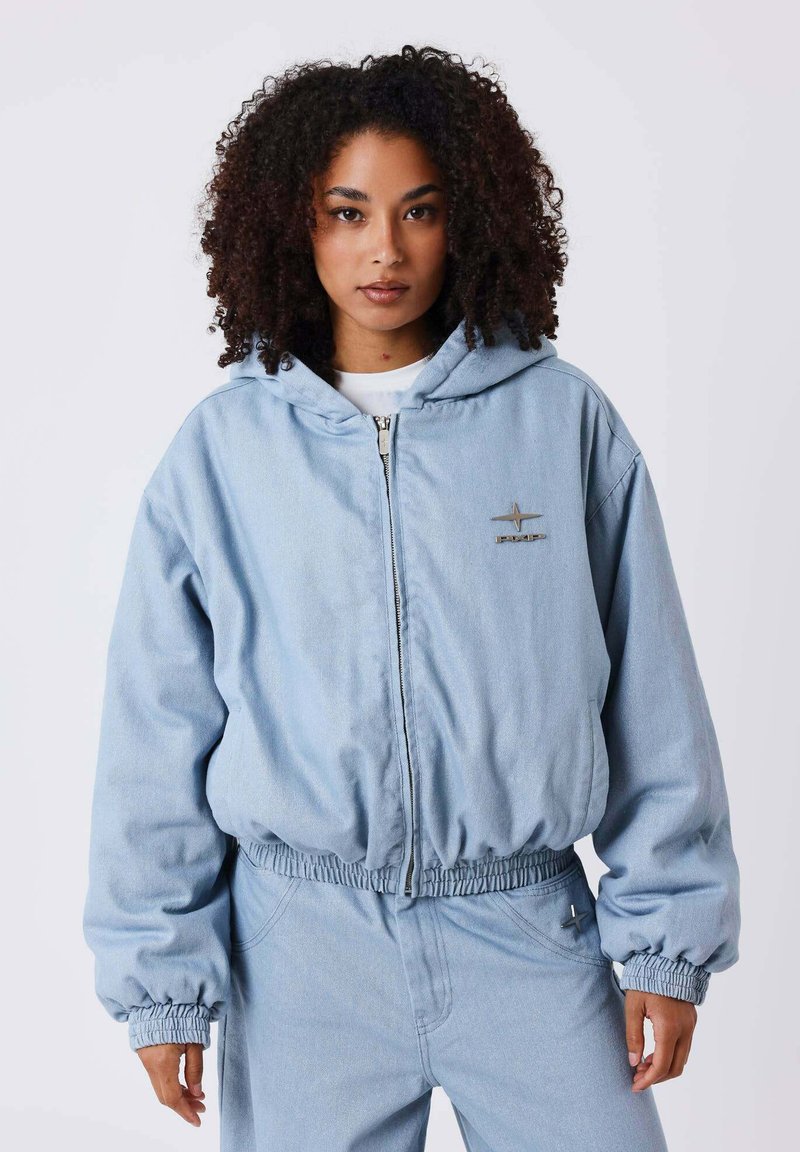 Femme aux cheveux bouclés portant un sweat à capuche bleu clair zippé et un pantalon assorti, debout devant un fond blanc uni.