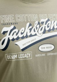 Beiger Stoff mit weißem und marineblauem Text, darunter "Fine Cotton Select", "Jack & Jones", "Nineteen Ninety", "Denim Legacy", "Raw Indigo" sowie die Städtenamen Barcelona und Kopenhagen.