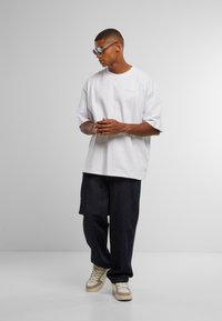 Bílý oversized tričko s krátkými rukávy, kombinované s tmavými džínovými kalhotami a béžovými teniskami. Doplňky zahrnují stříbrné sluneční brýle.