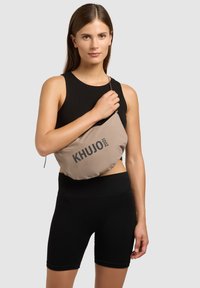 Femme en haut sans manches noir et short tenant un sac bandoulière beige avec le logo "KHUJO 2001" sur fond gris clair.