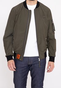 Olivgrön bomberjacka med dragkedja fram, ribbade ärmslut och en patch på vänster sida. Tillverkad av lätt, slätt material.