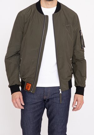 Giacca bomber verde oliva con zip frontale, polsini a coste e una toppa sul lato sinistro. Realizzata in materiale leggero e liscio.