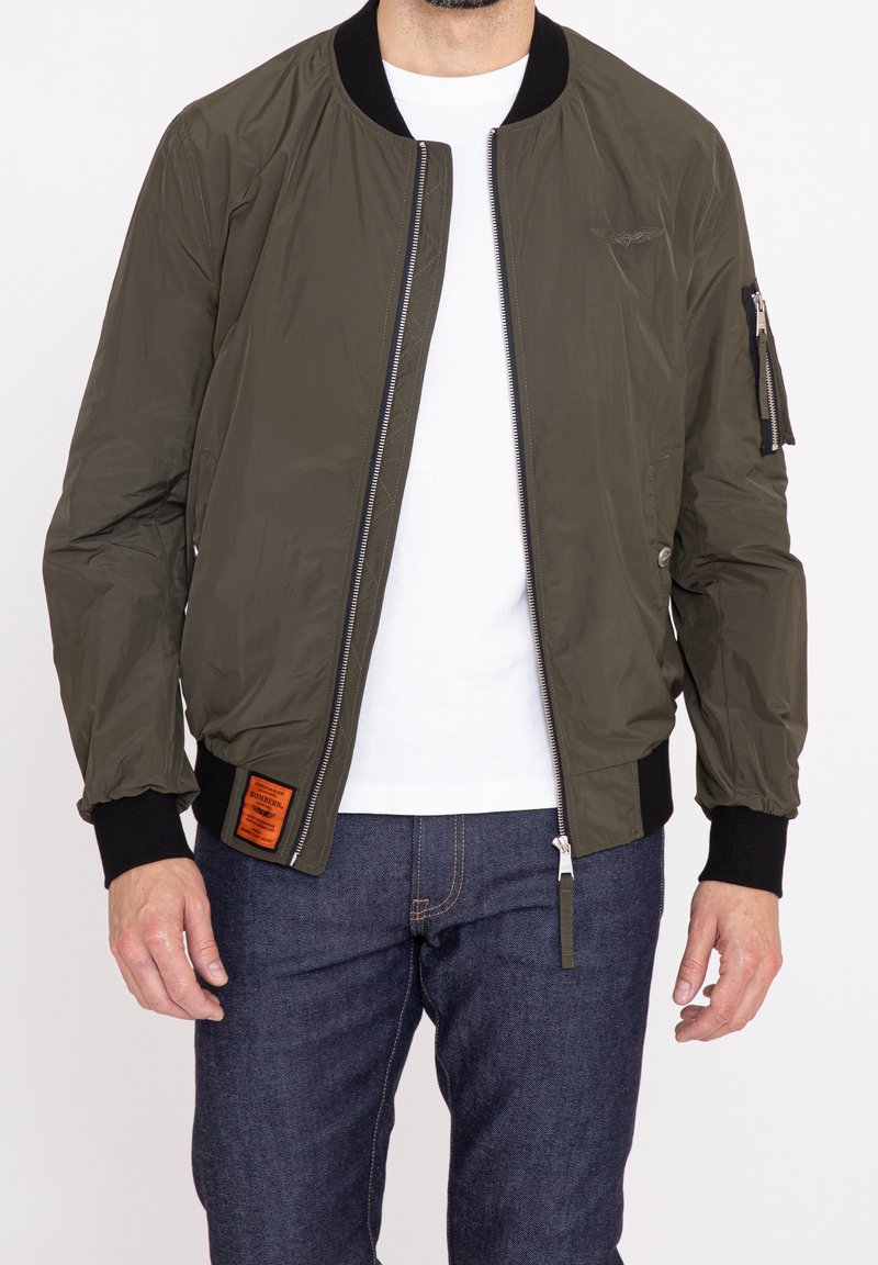 Olivgrön bomberjacka med dragkedja fram, ribbade ärmslut och en patch på vänster sida. Tillverkad av lätt, slätt material.