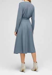 Kobieta w niebieskiej, midi sukience z długim rękawem i marszczonym stanikiem, stoi tyłem do kamery na szarym tle, w kremowych botkach za kostkę.