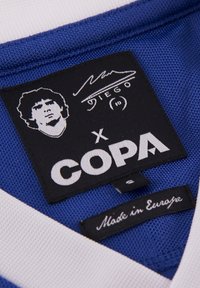 Cuello de camiseta deportiva azul con un ribete blanco, que presenta una etiqueta negra con un retrato gráfico, una firma y el nombre de la marca "COPA". Las etiquetas indican la talla y el origen.