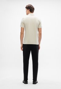 Polo shirt beige clair à manches courtes et col, associé à un pantalon noir et des chaussures sombres, vu de dos sur fond blanc.