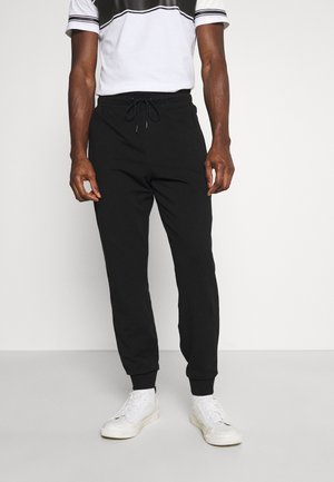 Mann in schwarzen Jogginghosen mit einem Kordelzug und weißen Sneakers, der vor einem schlichten hellen Hintergrund steht.