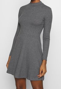 Robe grise côtelée à manches longues et col montant, avec un buste ajusté et une jupe évasée. Texture lisse et silhouette épurée.